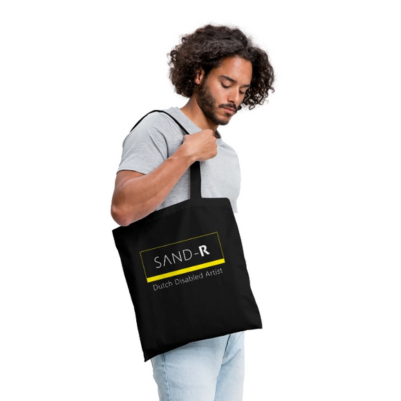 Mixed race man with dark curly hair, wearing a grey t-shirt and light blue jeans holding a black tote bag that is resting on his shoulder with text: SAND-R, Dutch Disabled Artist.